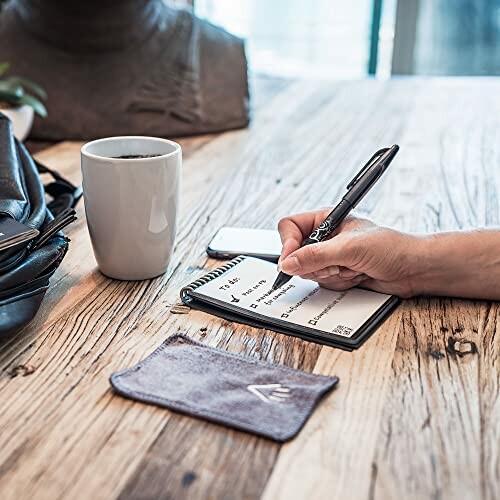 Person writing on a notepad beside a coffee mug and smartphone on a wooden table.