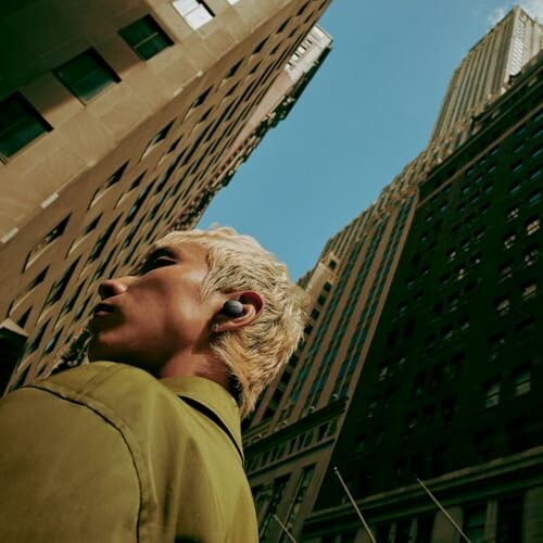 Person with blonde hair wearing earbuds, looking up at tall buildings in a city.